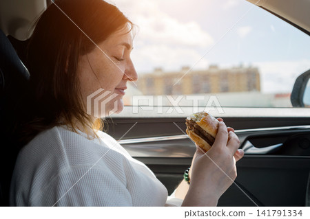 Woman enjoying a cheeseburger while traveling in car Woman enjoying a cheeseburger while traveling in car 141791334