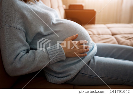 Pregnant woman resting on a sofa with hand on belly in a bright cozy living room Pregnant woman resting on a sofa with hand on belly in a bright cozy living room 141791642