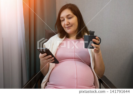 Pregnant woman relaxing with smartphone and mug in a cozy room Pregnant woman relaxing with smartphone and mug in a cozy room 141791644