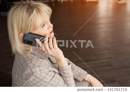 Mature blonde businesswoman in a plaid suit makes a focused phone call in an office, illustrating professional communication, remote work and confident business networking at daytime Mature blonde businesswoman in a plaid suit makes a focused phone call in an office, illustrating professional communication, remote work and confident business networking at daytime 141791719