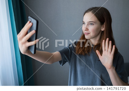 Young teenage girl smiles and waves at her smartphone during a casual video call at home, connecting with friends or family via mobile device for online conversation and greeting Young teenage girl smiles and waves at her smartphone during a casual video call at home, connecting with friends or family via mobile device for online conversation and greeting 141791720
