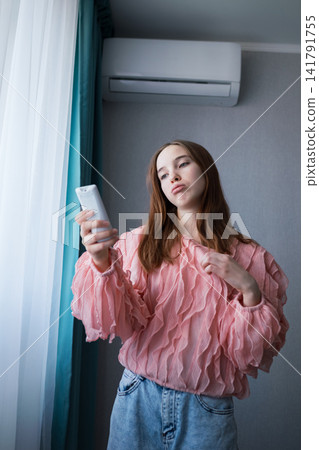 Young frustrated woman holding a remote control, operating the air conditioning unit indoors, seeking relief from hot temperature and feeling uncomfortable during summer Young frustrated woman holding a remote control, operating the air conditioning unit indoors, seeking relief from hot temperature and feeling uncomfortable during summer 141791755