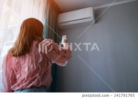 Woman adjusts split system air conditioner with remote control, cooling a sunny apartment for comfortable indoor climate and energy efficient relief during hot summer weather Woman adjusts split system air conditioner with remote control, cooling a sunny apartment for comfortable indoor climate and energy efficient relief during hot summer weather 141791765