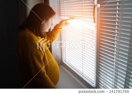 Pregnant woman standing by window blinds in warm morning light 141791789