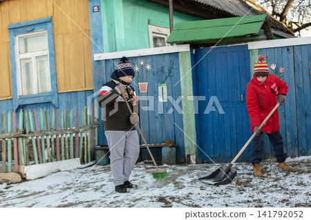 Two children shoveling snow in front of a colorful wooden house in winter attire Two children shoveling snow in front of a colorful wooden house in winter attire 141792052