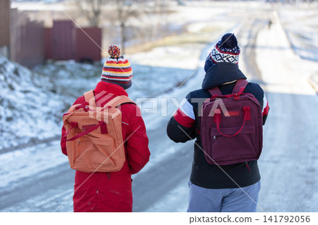 Two schoolboy boys in colorful winter hats and backpacks walk along a snowy road with their backs to the camera Two schoolboy boys in colorful winter hats and backpacks walk along a snowy road with their backs to the camera 141792056