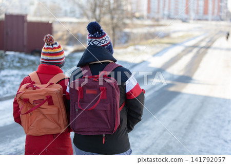 Schoolchildren with backpacks walk with their backs to the camera along a snowy road. Schoolchildren with backpacks walk with their backs to the camera along a snowy road. 141792057