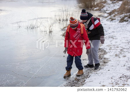 Two children dressed in bright winter clothes explore a snowy path near a frozen lake. They're trying to walk on dangerously thin ice Two children dressed in bright winter clothes explore a snowy path near a frozen lake. They're trying to walk on dangerously thin ice 141792062