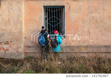 Two people with backpacks and winter hats looking into a barred window of an old weathered building Two people with backpacks and winter hats looking into a barred window of an old weathered building 141792067