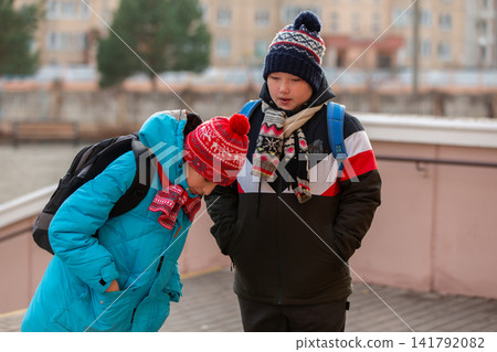 Kids enjoy time outdoors in winter, looking at something on the ground near a building by the river Kids enjoy time outdoors in winter, looking at something on the ground near a building by the river 141792082
