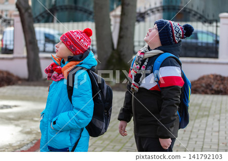 Kids look up at the sky while standing on a sidewalk near trees in winter outfit during daylight hours in a city park Kids look up at the sky while standing on a sidewalk near trees in winter outfit during daylight hours in a city park 141792103