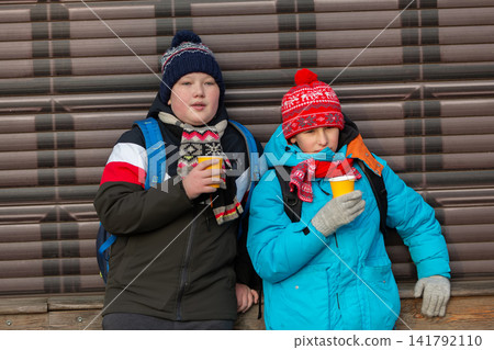 Two children drink warm beverages while leaning against a wall in a chilly outdoor setting during winter Two children drink warm beverages while leaning against a wall in a chilly outdoor setting during winter 141792110