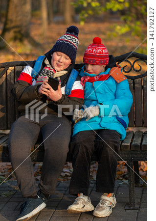 Two school age boys sitting on a bench and looking at a smartphone in the park Two school age boys sitting on a bench and looking at a smartphone in the park 141792127