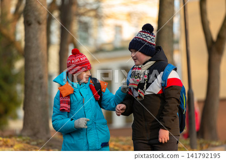Two schoolboy boys with backpacks walk in an autumn park in bright jackets and hats, and leaves are falling around them. Two schoolboy boys with backpacks walk in an autumn park in bright jackets and hats, and leaves are falling around them. 141792195