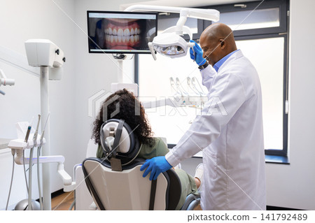 Black male dentist preparing examination light for female patient in dental clinic Black male dentist preparing examination light for female patient in dental clinic 141792489