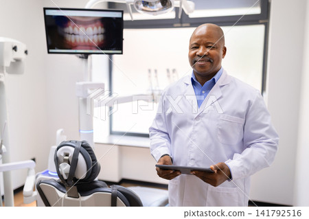 Black male dentist holding digital tablet beside dental chair in modern clinic Black male dentist holding digital tablet beside dental chair in modern clinic 141792516