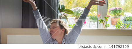 A middle-aged woman sits up in bed, stretching her arms above her head. She looks tired and has not had enough sleep. The room is bright with natural light and filled with plants, banner A middle-aged woman sits up in bed, stretching her arms above her head. She looks tired and has not had enough sleep. The room is bright with natural light and filled with plants, banner 141792599