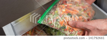 A man reaches into the refrigerator and takes out a bag of frozen vegetables. He is preparing to cook during the afternoon in a kitchen setting, banner A man reaches into the refrigerator and takes out a bag of frozen vegetables. He is preparing to cook during the afternoon in a kitchen setting, banner 141792681