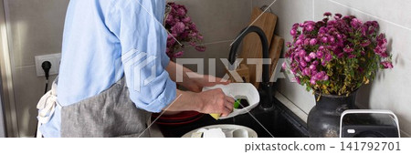 A man stands at the sink in his kitchen, cleaning dirty dishes and utensils. Flowers are placed on the counter, adding color to the scene in daylight, banner A man stands at the sink in his kitchen, cleaning dirty dishes and utensils. Flowers are placed on the counter, adding color to the scene in daylight, banner 141792701