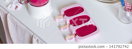 In a kitchen, a blender is mixing strawberries, bananas, and beets. Ice cream molds sit on the table filled with the mixture. A ripe banana and a beet are nearby, banner In a kitchen, a blender is mixing strawberries, bananas, and beets. Ice cream molds sit on the table filled with the mixture. A ripe banana and a beet are nearby, banner 141792742