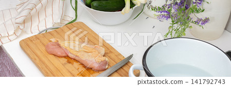 A person is cutting a chicken breast on a wooden board in a kitchen. There are green onions and cucumbers in a bowl nearby. A pot with water is on the stove ready for cooking, banner A person is cutting a chicken breast on a wooden board in a kitchen. There are green onions and cucumbers in a bowl nearby. A pot with water is on the stove ready for cooking, banner 141792743