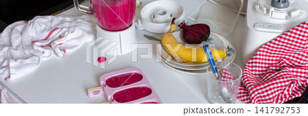 In a kitchen, a blender is mixing strawberries, bananas, and beets. Ice cream molds sit on the table filled with the mixture. A ripe banana and a beet are nearby, banner In a kitchen, a blender is mixing strawberries, bananas, and beets. Ice cream molds sit on the table filled with the mixture. A ripe banana and a beet are nearby, banner 141792753
