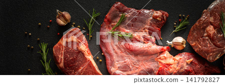 Different cuts of beef steak are arranged on a black surface. Fresh herbs, spices, and garlic accompany the meat, ready for cooking or grilling at a kitchen or market, banner Different cuts of beef steak are arranged on a black surface. Fresh herbs, spices, and garlic accompany the meat, ready for cooking or grilling at a kitchen or market, banner 141792827