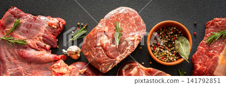 Several types of beef steak are laid out on a dark surface with fresh herbs and spices. Ingredients include garlic, pepper, and oil, showing preparation for cooking in a kitchen, banner Several types of beef steak are laid out on a dark surface with fresh herbs and spices. Ingredients include garlic, pepper, and oil, showing preparation for cooking in a kitchen, banner 141792851