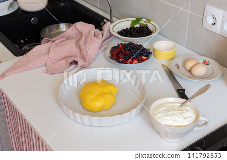 A person prepares ingredients for a strawberry, currant, and blueberry shortbread pie with sour cream sauce on a kitchen counter. Fresh fruits and dough are arranged in bowls and plates A person prepares ingredients for a strawberry, currant, and blueberry shortbread pie with sour cream sauce on a kitchen counter. Fresh fruits and dough are arranged in bowls and plates 141792853