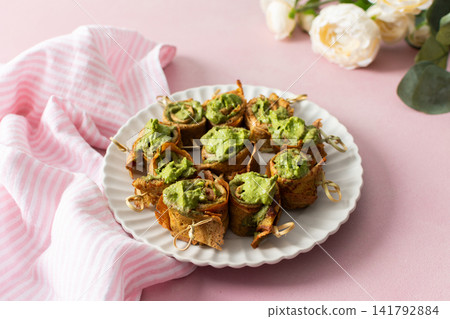 Plate holds pesto pancake rolls topped with green spread and garnished with fresh lemon slices. A bowl of matching green sauce sits alongside on a simple set up Plate holds pesto pancake rolls topped with green spread and garnished with fresh lemon slices. A bowl of matching green sauce sits alongside on a simple set up 141792884