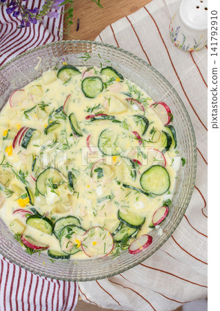 A bowl of okroshka filled with potatoes, chicken breast, cucumbers, herbs, and boiled eggs sits on a table. Nearby, a floral vase adds color to the setting A bowl of okroshka filled with potatoes, chicken breast, cucumbers, herbs, and boiled eggs sits on a table. Nearby, a floral vase adds color to the setting 141792910
