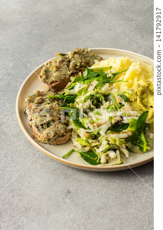 A plate shows a serving of boiled potatoes with hummus, a gluten free bread sandwich filled with herring mince, and fresh salad. This breakfast offers a nutritious start to the day 141792917