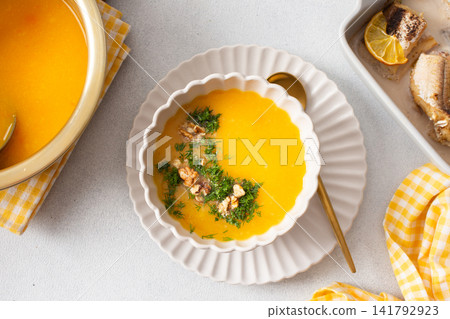 Pumpkin cream soup sits in a bowl with toppings next to a dish of baked hake and lemon. A spoon rests beside the soup in a clean dining area Pumpkin cream soup sits in a bowl with toppings next to a dish of baked hake and lemon. A spoon rests beside the soup in a clean dining area 141792923