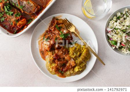 Plates hold amaranth, millet, and quinoa porridge served with baked white hake in tomato sauce. Fresh salad is beside the porridge. A bowl of kiwi sits nearby for dessert 141792971