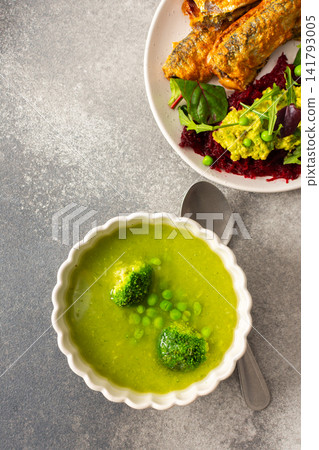 Cream of green pea and broccoli soup is served in a bowl with green peas on top. Baked Balkan herring and boiled beets with hummus are on a plate beside the soup Cream of green pea and broccoli soup is served in a bowl with green peas on top. Baked Balkan herring and boiled beets with hummus are on a plate beside the soup 141793005
