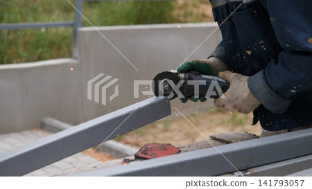 Close view of worker using handheld angle grinder on steel beam at outdoor construction site, sparks scattering from metal frame during fabrication and repair work in soft daylight Close view of worker using handheld angle grinder on steel beam at outdoor construction site, sparks scattering from metal frame during fabrication and repair work in soft daylight 141793057