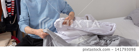 A man aged 53 is in his bedroom folding and changing bed linens. He sits on the bed with freshly cleaned sheets and a duvet beside him, focused on the task at hand, banner A man aged 53 is in his bedroom folding and changing bed linens. He sits on the bed with freshly cleaned sheets and a duvet beside him, focused on the task at hand, banner 141793098