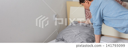 A 53-year-old man is replacing the bed linen in his bedroom. He is focused on his task, arranging pillows and sheets neatly. Sunlight comes into the room, which has plants and simple decor, banner A 53-year-old man is replacing the bed linen in his bedroom. He is focused on his task, arranging pillows and sheets neatly. Sunlight comes into the room, which has plants and simple decor, banner 141793099