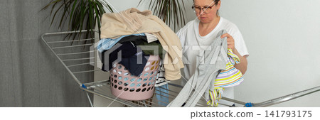 A woman is taking laundry out of the dryer and putting it into a basket. She is in a simple room with plants nearby. Her focus is on the task at hand, banner A woman is taking laundry out of the dryer and putting it into a basket. She is in a simple room with plants nearby. Her focus is on the task at hand, banner 141793175