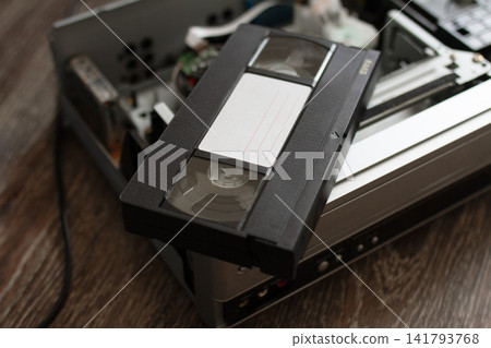 Black retro VHS video cassette tape with blank white label lying on top of disassembled, broken or dusty old video player mechanism, nostalgic analog media equipment Black retro VHS video cassette tape with blank white label lying on top of disassembled, broken or dusty old video player mechanism, nostalgic analog media equipment 141793768