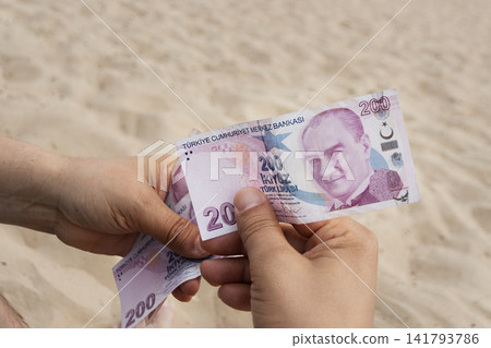 Close up of person hands holding and counting two hundred Turkish Lira bills on sandy beach background. Concept of travel budget, vacation spending, and Turkish currency Close up of person hands holding and counting two hundred Turkish Lira bills on sandy beach background. Concept of travel budget, vacation spending, and Turkish currency 141793786