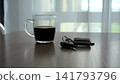 A Close-Up View of a Glass of Coffee Next to Car Keys on a Wooden Table with Soft Natural Light Filtering Through Sheer Curtains. 141793796