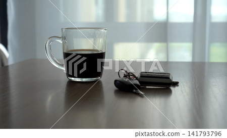 A Close-Up View of a Glass of Coffee Next to Car Keys on a Wooden Table with Soft Natural Light Filtering Through Sheer Curtains. A Close-Up View of a Glass of Coffee Next to Car Keys on a Wooden Table with Soft Natural Light Filtering Through Sheer Curtains. 141793796