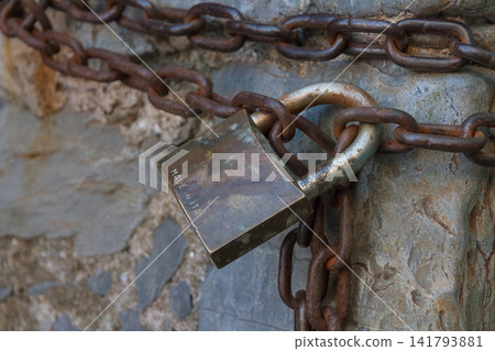 Close up view of weathered vintage brass padlock made in Italy locking heavy rusty iron chains. Set against an ancient rough texture stone wall, creating a strong visual concept for security, safety, Close up view of weathered vintage brass padlock made in Italy locking heavy rusty iron chains. Set against an ancient rough texture stone wall, creating a strong visual concept for security, safety, 141793881