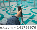 First person POV view of hand in denim jacket holding black rubber handle of outdoor gym equipment on turquoise rubber sports ground background 141793976