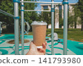 Close up view of human hand holding craft paper takeaway coffee cup with transparent plastic lid against blurry outdoor workout gym and turquoise sports ground 141793980
