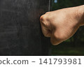 Close up side view of man bare fist punching heavy black leather boxing bag outdoors on sunny day 141793981