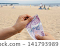 Close up of person hands holding three 200 Turkish lira banknotes in hands against blurry sandy beach with people and sea background 141793989
