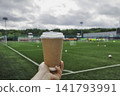 Close up of human hand holding craft paper takeaway coffee cup with white plastic lid against green soccer football field on cloudy day 141793991