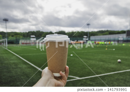 Close up of human hand holding craft paper takeaway coffee cup with white plastic lid against green soccer football field on cloudy day 141793991